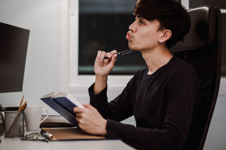 Young handsome asian man thinking new idea from reading book at work desk late at night, Knowledge...