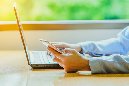 Businessman working with smart phone and laptop computer on wooden desk ; remote working concept; home business concept.の写真素材