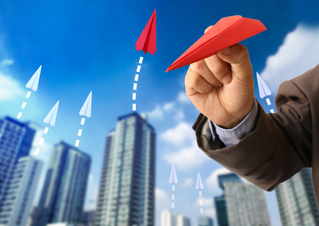 Business man in suit holding red paper airplane gesture to throwing with blurred image of building background and blue sky, Target on business concept.の写真素材