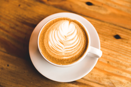 A carefully prepared cappuccino in a simple white cup and plate, sitting on wooden table. Leaf design in cream on top.の写真素材
