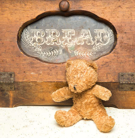 Small brown teddy bear seated in front of a older looking brown wooden bread box.の写真素材