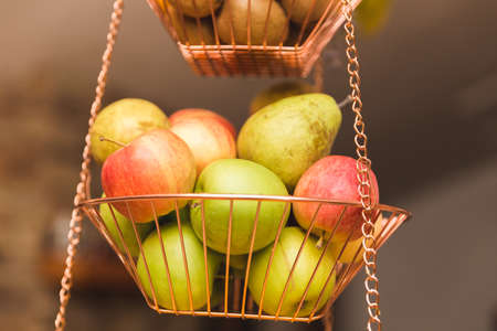 Red and green apples with pears in a hanging metal basket inside a kitchen.の写真素材