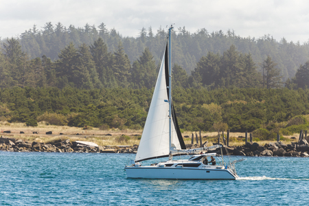 Sailboat sailing through a body of blue water lined by a forest and rocky shore.の写真素材