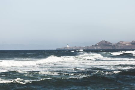 Ocean waves beginning to break near the coastline with a lighthouse on a rocky outcropping in the distance.の写真素材