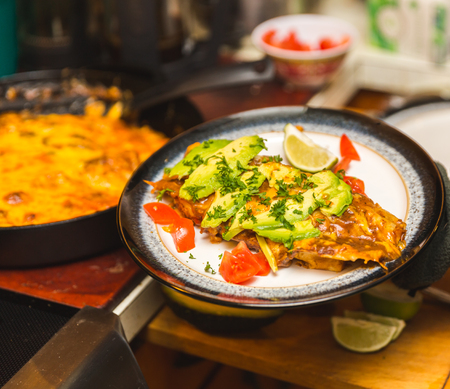 Burrito or enchilada on plate in kitchen setting. Ingredients include tortilla, cheese, peppers, avocados, tomatoes, and lime.の写真素材