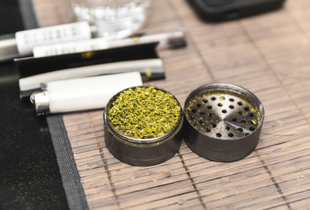Ground cannabis inside a steel grinder that's been opened and set on a table placemat. Lighter and other marijuana-related items can be seen in background.の写真素材
