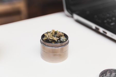 Steel grinder with green cannabis flower (buds) inside. Laptop in background.の写真素材