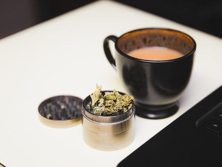 Cannabis or marijuana flower in a steel grinder on a desk, next to a cup of coffee and a laptop.の写真素材