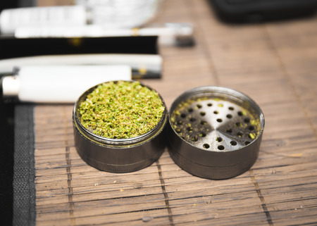Ground cannabis inside a steel grinder that's been opened and set on a table placemat. Lighter and other marijuana-related items can be seen in background.の写真素材