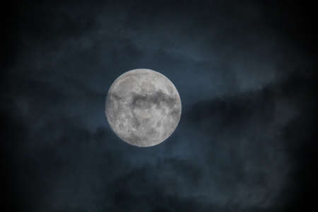 Full moon captured behind clouds in night sky. Dark clouds cover a bright white full moon. Captured...