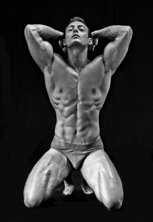 Black and white shot of young bodybuilder kneeling with arms up, isolated on blackの写真素材