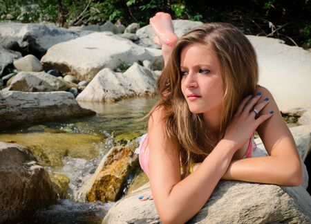 Pretty brunette girl in bikini laying on rocks by small water streamの写真素材
