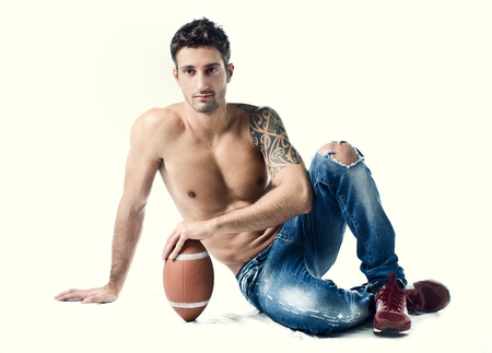 Athletic young man shirtless wearing jeans, with american football ball sitting on floor, isolatedの写真素材