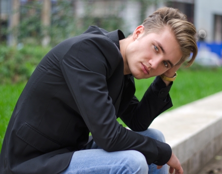 Serious or sad blond young man in jeans and jacket, sitting outdoors looking in cameraの写真素材