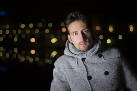Attractive young man portrait at night with city lights behind him, looking at cameraの写真素材