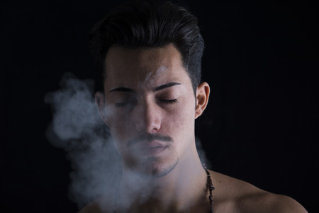 Attractive young man smoking cigarette, eyes closed, isolated on black backgroundの写真素材