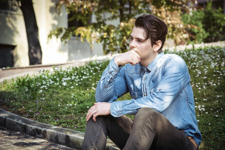 Young man sitting outdoors in public park, with hand on his chin looking away thinkingの写真素材