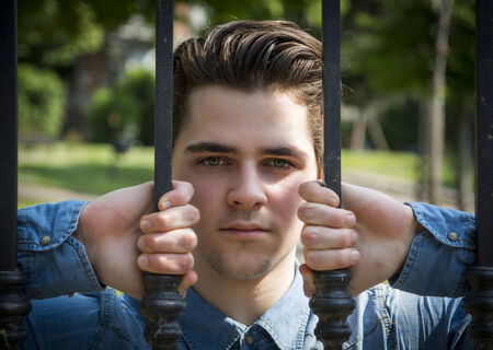 Attractive young man behind metal gate, gripping bars with his handsの写真素材