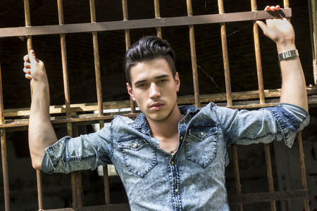 Handsome black haired young man in denim shirt against metal cage barsの写真素材