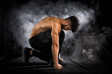 Handsome shirtless muscular young man kneeling down on black background, smoke around him. Looking downの写真素材