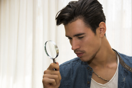 Young man looking through a magnifying glass in a conceptual image of a detective searching for clues, inspection, examination or analysisの写真素材