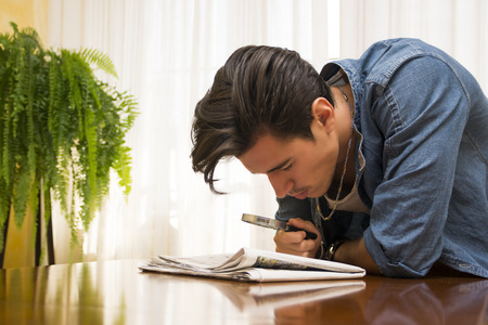 Young man bending over a table at home reading a newspaper with a magnifying glassの写真素材