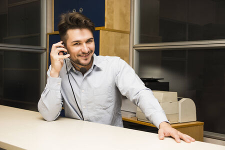 Businessman or customer (support) service man taking a call on his headset phone as he sits at a table in the office looking up to smile at the camera as he listens to the conversationの写真素材