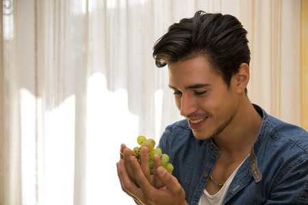 Young man holding and looking at a large delicious ripe bunch of grapes in his hands, smilingの写真素材