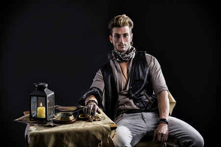 Good Looking Young Man in Pirate Fashion Outfit Sitting next to Table with Candle Lamp, Compass, Goldの写真素材
