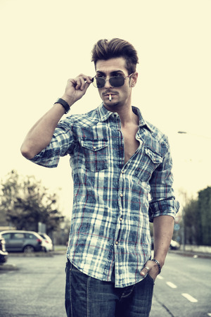 Trendy cool young man standing outside smoking, lowering sunglasses, wearing shirt and jeansの写真素材