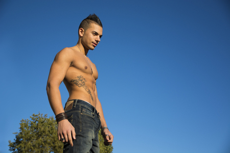 Handsome fit athletic shirtless young man against blue sky shot from below, looking at cameraの写真素材