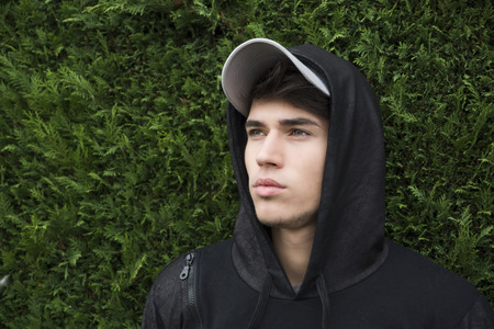 Attractive young man with hoodie and baseball cap against green bush "wall", sad or thinkingの写真素材