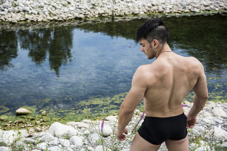 Back of muscular shirtless young man outdoor near river or lake, looking down to a sideの写真素材