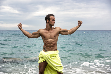 Attractive shirtless muscleman on the beach covering with towel in muscular poseの写真素材