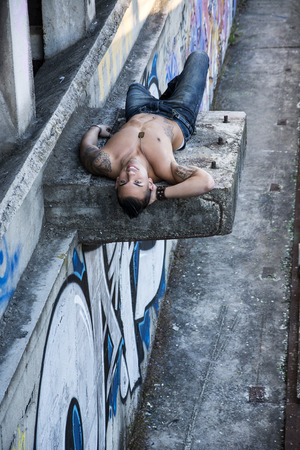 Handsome shirtless young man laying down on concrete structure above the ground, smiling at cameraの写真素材