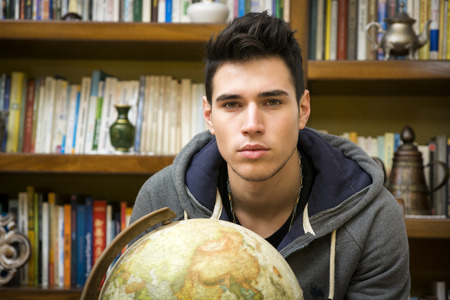 Handsome young man holding a globe indoors at home looking at the camera in front of a bookcase filled with books in a conceptual imageの写真素材