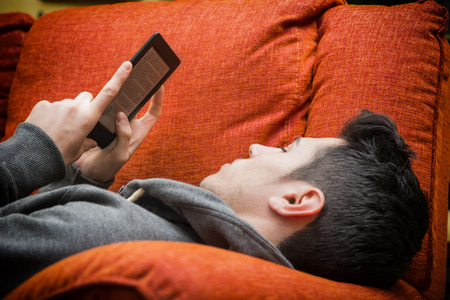 Handsome young man at home reading with ebook reader lying on a couchの写真素材