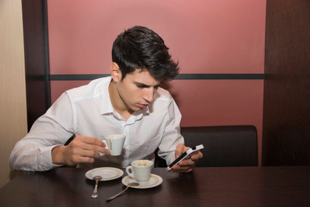 Close up Handsome Young Man Drinking Coffee at the Shop While Looking at Mobile Phoneの写真素材