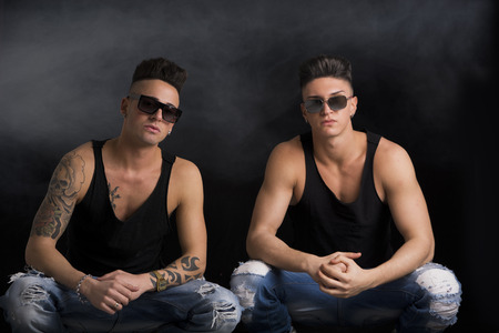 Two hip trendy male friends in studio shot sitting on the floor, wearing sunglasses and black tanktopsの写真素材
