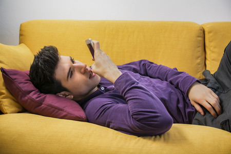 Attractive young man recording voice message on cell phone while laying on couch, with serious expressionの写真素材