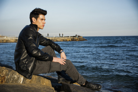 Handsome fashionable young man at the seaside along the shore overlooking the ocean or sea with his black leather jacket looking at camera, large copyspaceの写真素材
