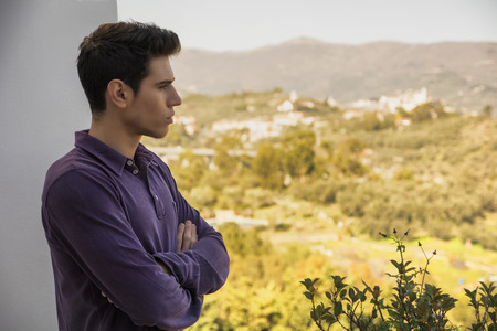 Attractive young man standing with folded arms looking over a rural landscape with a distant village with a serious expressionの写真素材