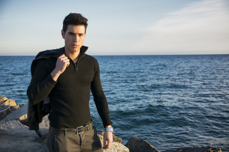 Handsome fashionable young man at the seaside walking along a promenade overlooking the ocean with his jacket over his shoulder looking at the cameraの写真素材