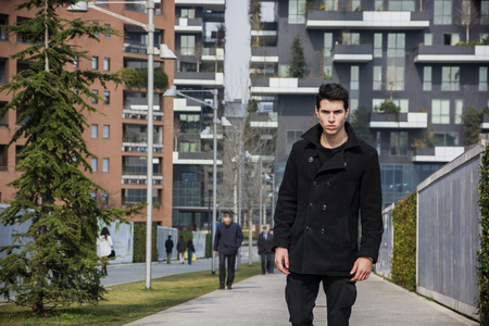 Stylish Young Handsome Man in Black Coat Standing in City Center Street with Skyscraper Behind Him, Looking to the Cameraの写真素材