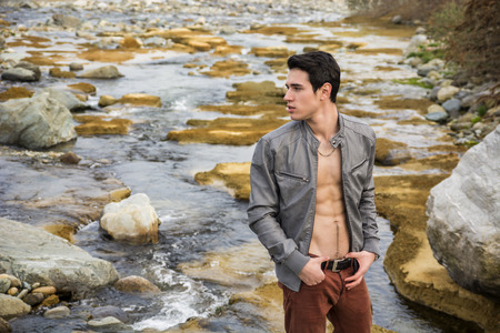 Handsome fit shirtless young man next to water pond or river, wearing grey leather jacket open on naked torsoの写真素材