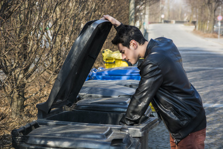 Attractive young man putting out rubbish standing with the lid up on a bin in a row of garbage bins...