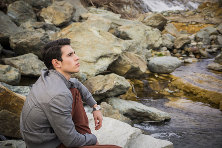 Handsome young man outdoor sitting alone at river or water stream looking up thinkingの写真素材