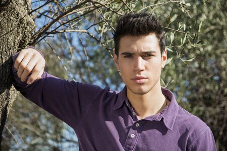 Handsome young man leaning against olive tree, looking at camera, in a sunny dayの写真素材