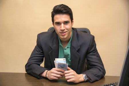 Close up Smiling Young Businessman, Sitting at his Worktable, Counting Cash on Handの写真素材
