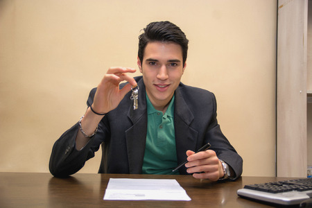 Close up Young Real Estate Broker Showing a Key While Sitting at his Table and Looking at the Camera.の写真素材